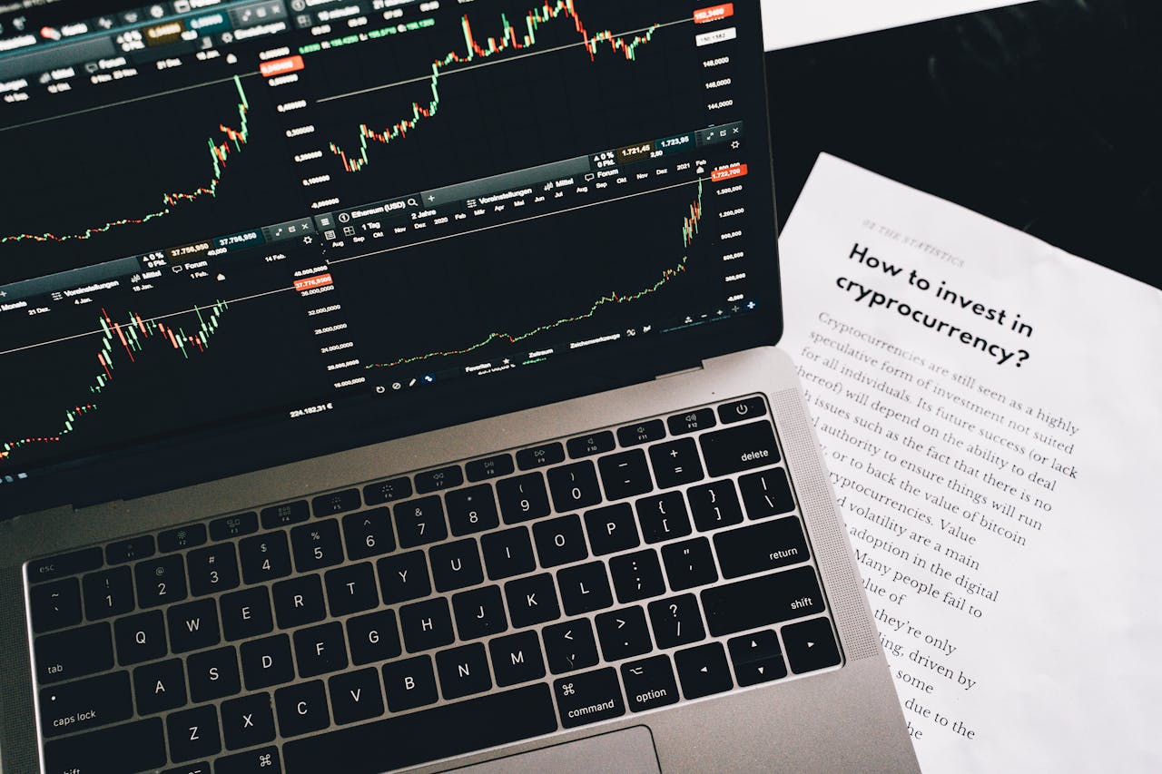 Close-up of a laptop displaying stock market graphs with a paper on investing in cryptocurrency, indoors.